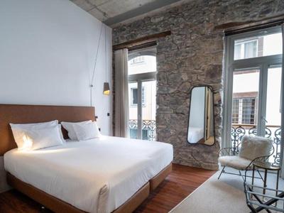 Hell eingerichtetes Schlafzimmer mit Steinwand, großem Bett, zwei Fenstern und kleinem Spiegel.