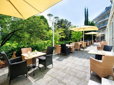 Outdoor hotel terrace with tables, chairs, umbrellas, and greenery overlooking a building.