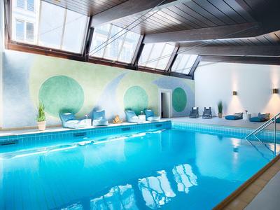 Indoor pool with modern seating and wall art under a sloped ceiling with large windows.