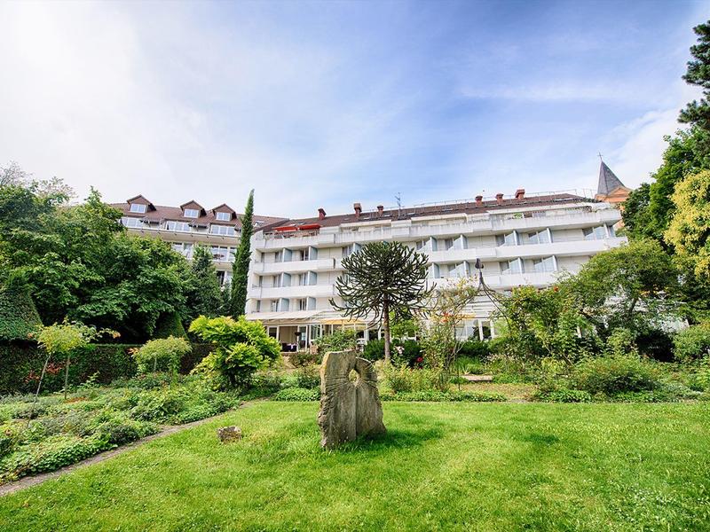 Large hotel building with white balconies behind lush green garden and trees under blue sky.