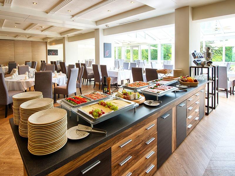 Bright hotel buffet area with plates, salads, and chairs near large windows