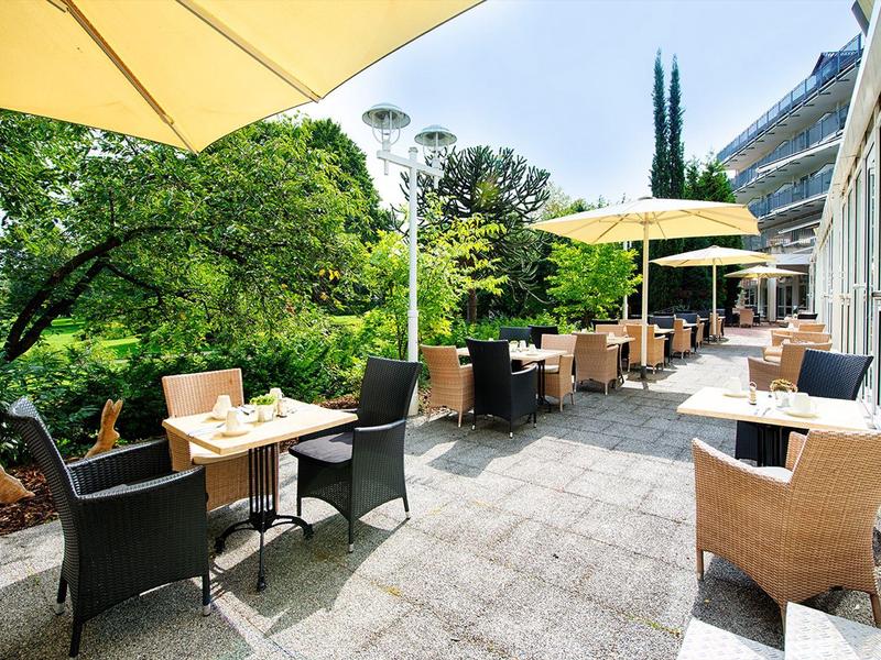 Outdoor hotel terrace with tables, chairs, umbrellas, and greenery overlooking a building.