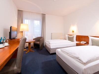 Helles Hotelzimmer mit zwei Einzelbetten, Schreibtisch, Sesseln und Fenster mit Vorhängen.