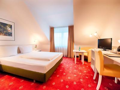 Hell eingerichtetes Hotelzimmer mit Doppelbett, Schreibtisch und rotem Teppichboden.