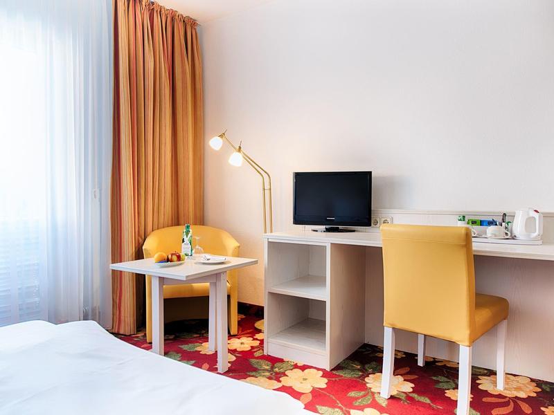 Helles Hotelzimmer mit orangem Teppich, gelbem Stuhl, Schreibtisch, TV und Vorhängen am Fenster.