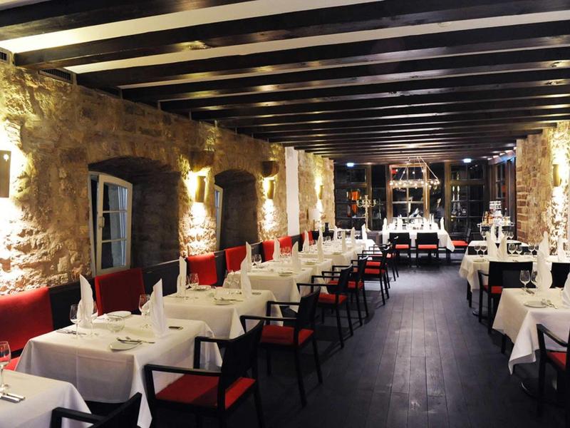 Eleganter Gastraum mit weißen Tischdecken, roten Sitzpolstern, Steinwänden und Holzdeckenbalken.