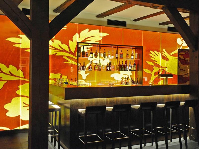 Moderner Barbereich mit dunklem Holztresen und hohen Stühlen vor einer Wand mit orange-gelben Blättermotiven.