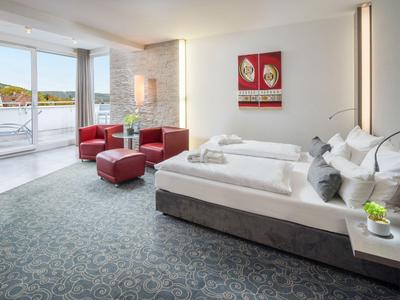 Helles Hotelzimmer mit Doppelbett, rotem Sitzbereich und Balkon mit Blick auf das Wasser.