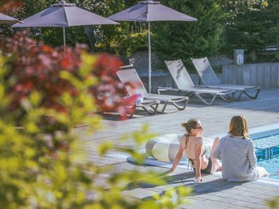 Due persone sono sedute a bordo piscina vicino a lettini e ombrelloni in un ambiente verde.