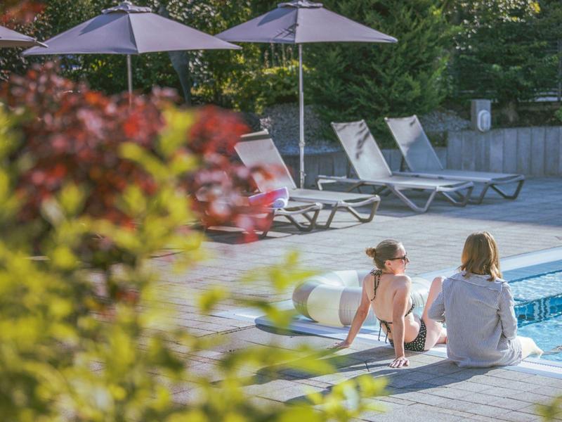 Due persone sono sedute a bordo piscina vicino a lettini e ombrelloni in un ambiente verde.
