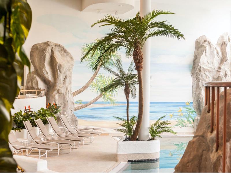 Zona piscina con lettini e palme davanti a una parete dipinta con una scena marina.