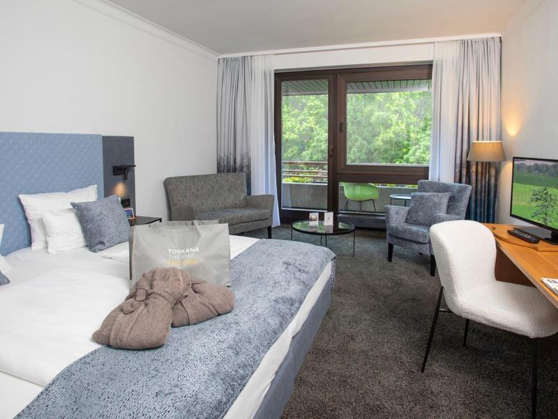 Nowoczesny pokój hotelowy z dużym łóżkiem, częścią wypoczynkową i biurkiem przy oknie z widokiem na zieleń.