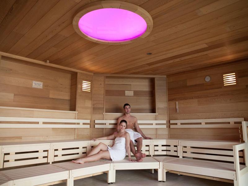 Przytulna sauna z drewnianymi panelami, ławkami i okrągłym różowym światłem sufitowym.