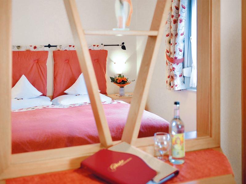 Schlafzimmer mit rotem Bett, Kissen, Buch, Wasserflasche und Fenster mit Vorhängen.