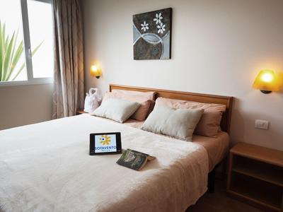 Hell eingerichtetes Schlafzimmer mit Doppelbett, zwei Nachttischen, Bild an der Wand und Tablet auf der Decke.
