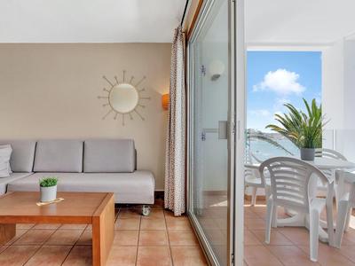 Gemütliche Wohnzimmer-Ecke mit hellgrauer Couch, Holztisch und Zugang zu Balkon mit weißem Tisch und Stühlen.