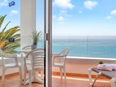 Balkon mit weißen Stühlen, Tisch, Liegestuhl und Meerblick unter blauem Himmel.