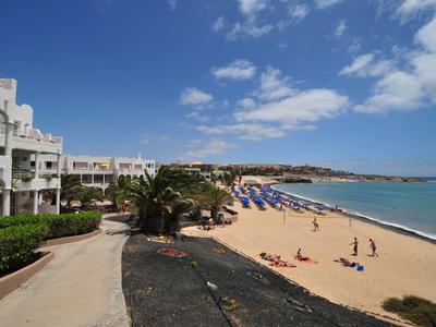 Strand mit hellem Sand, blauer Himmel, einigen Wolken, Hotelgebäuden links und Sonnenschirmen am Meer.