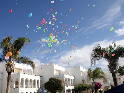 Bunte Luftballons steigen über weißen Häusern und Palmen an einem sonnigen Himmel empor.