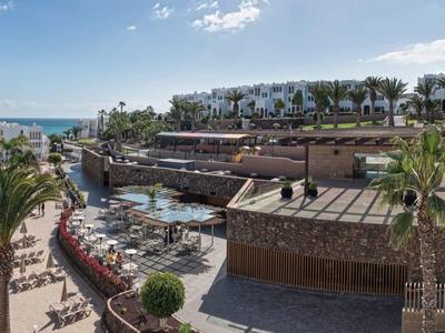 Moderner Strandkomplex mit Pool, Palmen, Holzterrassen und Blick auf Meer und bewölkten Himmel.