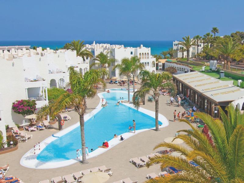 Hotelanlage mit einem großen Pool, weißen Gebäuden und Palmen, nahe dem Meer.