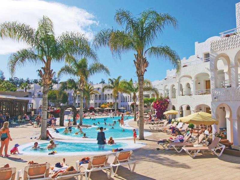 Belebter Poolbereich mit Palmen und weißer Hotelarchitektur bei sonnigem Himmel.