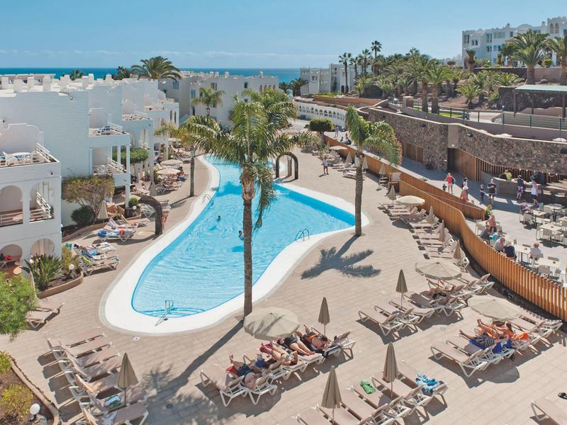 Poollandschaft mit Liegestühlen und weißen Gebäuden in einem Urlaubsresort am Meer