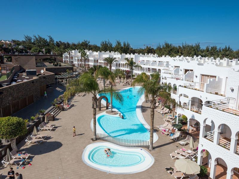 Hotelkomplex mit Pool und Liegestühlen in sonniger Lage mit weißen Gebäuden und Palmen.