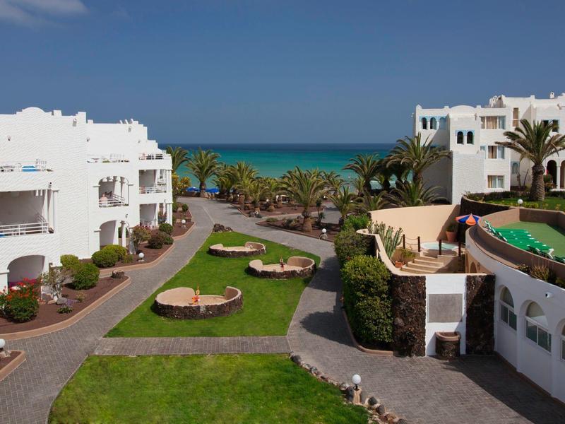 Blick auf Hotelanlage mit weißen Gebäuden, Palmen und Meer im Hintergrund.