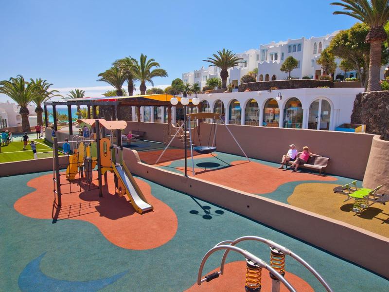 Bunter Kinderspielplatz mit Rutschen und Schaukeln vor einem Hotel mit Palmen.