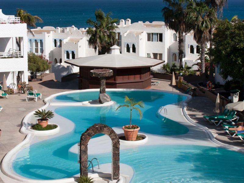 Beheizter Pool mit Liegestühlen und weißen Gebäuden am Meer bei klarem Himmel.