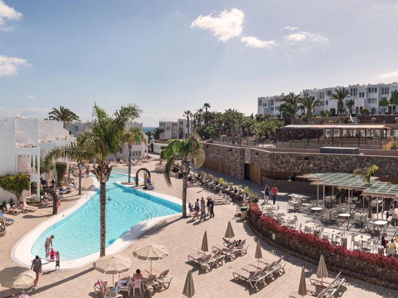 Ein Resort mit einem Pool, Liegestühlen und einem großen Gastronomiebereich im Freien bei sonnigem Wetter.