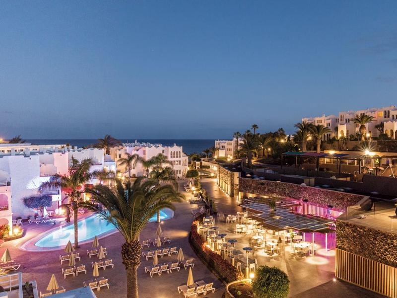 Abendaufnahme eines beleuchteten Hotelkomplexes mit Pools und Meer im Hintergrund.