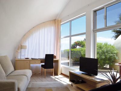 Helles Wohnzimmer mit Sofa, Schreibtisch und TV vor großen Fenstern mit Blick ins Grüne.