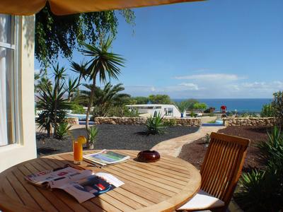 Blick von einer überdachten Terrasse mit Holztisch und Stuhl auf Palmen, Pool und Meer im Hintergrund.