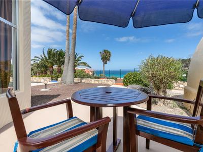 Terrasse mit rundem Holztisch und zwei Stühlen mit blau gestreiften Polstern, Blick aufs Meer.