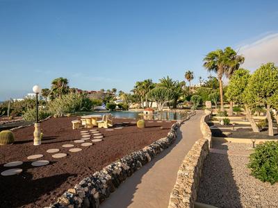 Ein gepflegter Garten mit Palmen, Sitzbereichen und einem Weg unter blauem Himmel.