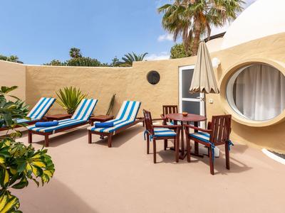 Sonnige Terrasse mit blauen Liegestühlen, Tisch und Stühlen vor einem runden Fenster.