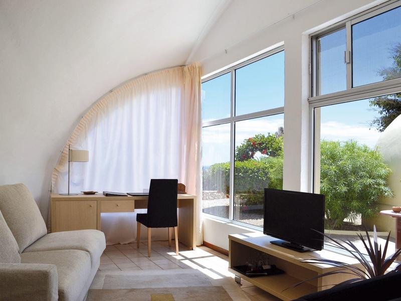 Helles Wohnzimmer mit Sofa, Schreibtisch und TV vor großen Fenstern mit Blick ins Grüne.