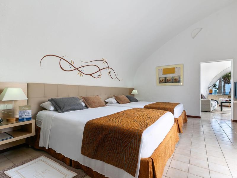 Helles Hotelzimmer mit zwei Einzelbetten, brauner Bettdecke, modernem Wanddekor und Blick in angrenzenden Raum.