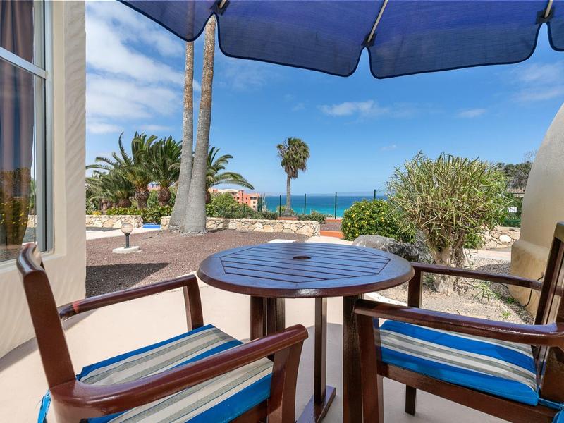 Terrasse mit rundem Holztisch und zwei Stühlen mit blau gestreiften Polstern, Blick aufs Meer.