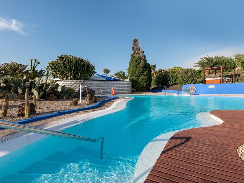 Freizeitpool mit Holzterrasse, Sonnenschirmen und Palmen unter blauem Himmel.