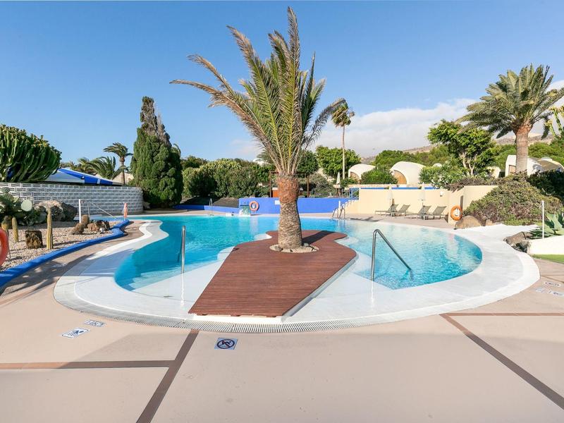 Runder Pool mit Palme in der Mitte, umgeben von Liegestühlen und Sonnenschirmen.