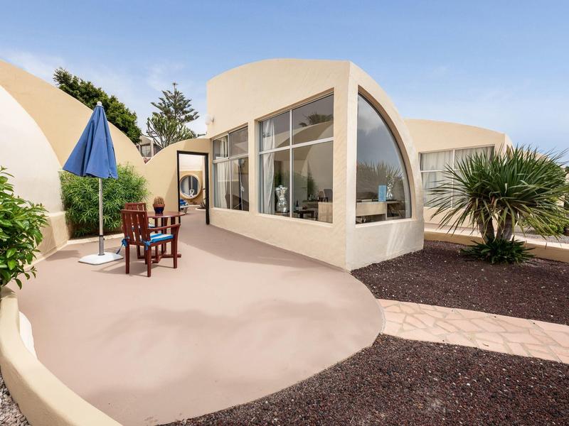 Moderner runder Bungalow mit Terrasse, Stühlen und blauem Sonnenschirm in sonniger Landschaft.