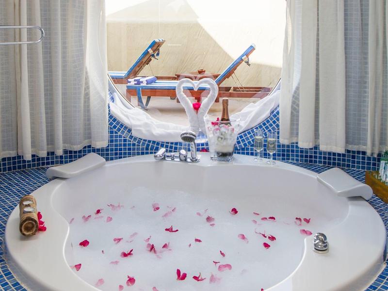 Runde Badewanne mit Schaum und Rosenblättern, vor rundem Fenster mit Sicht auf Sonnenschirme.
