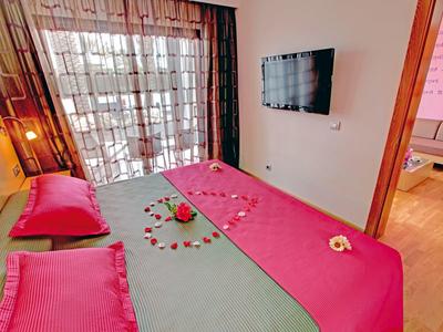 Gemütliches Schlafzimmer mit großem Bett, roten Decken, Kissen und Herz aus Rosenblättern.