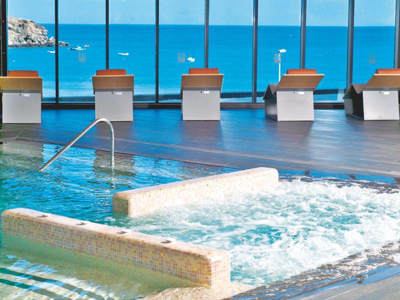 Overdekt zwembad met jacuzzi en panoramische ramen met uitzicht op zee en boten.