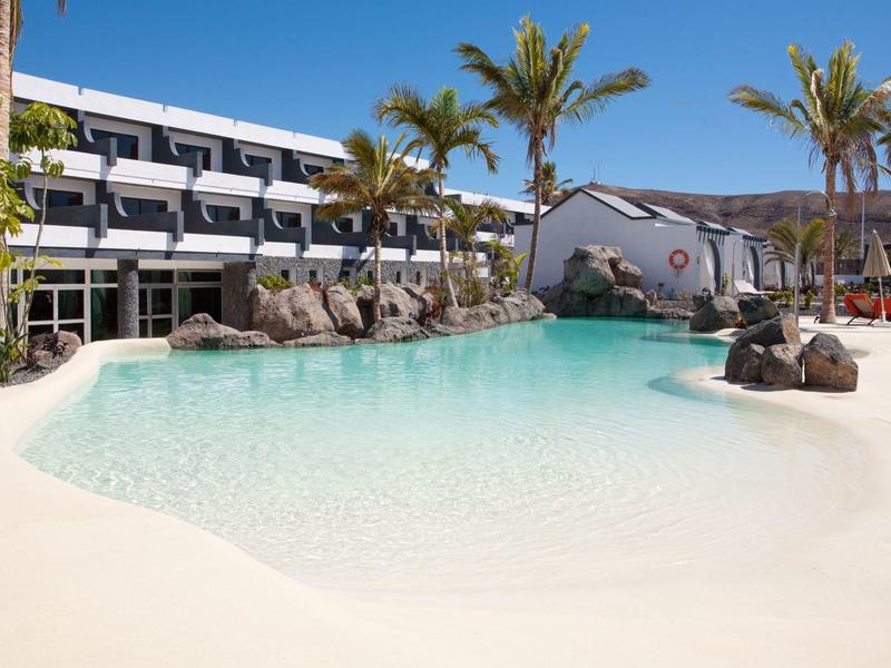 Modern hotelzwembad met helder water, witte zandvlakten en palmbomen onder een blauwe lucht.