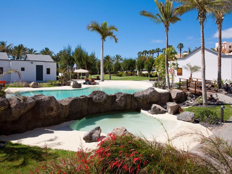 Hotelzwembad met rotsen, palmbomen en omliggende gebouwen onder blauwe lucht