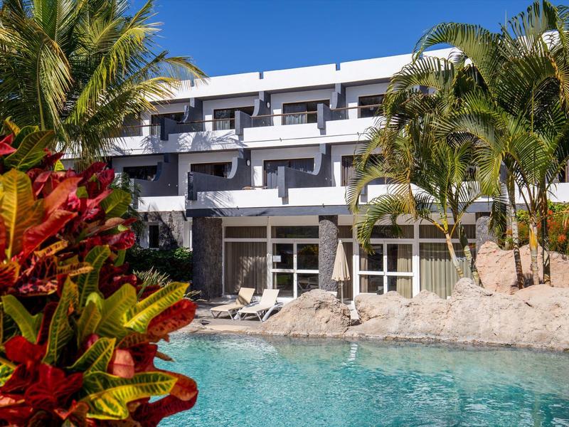 Hotelgebouw met palmbomen en zwembad op de voorgrond, omgeven door tropische planten.